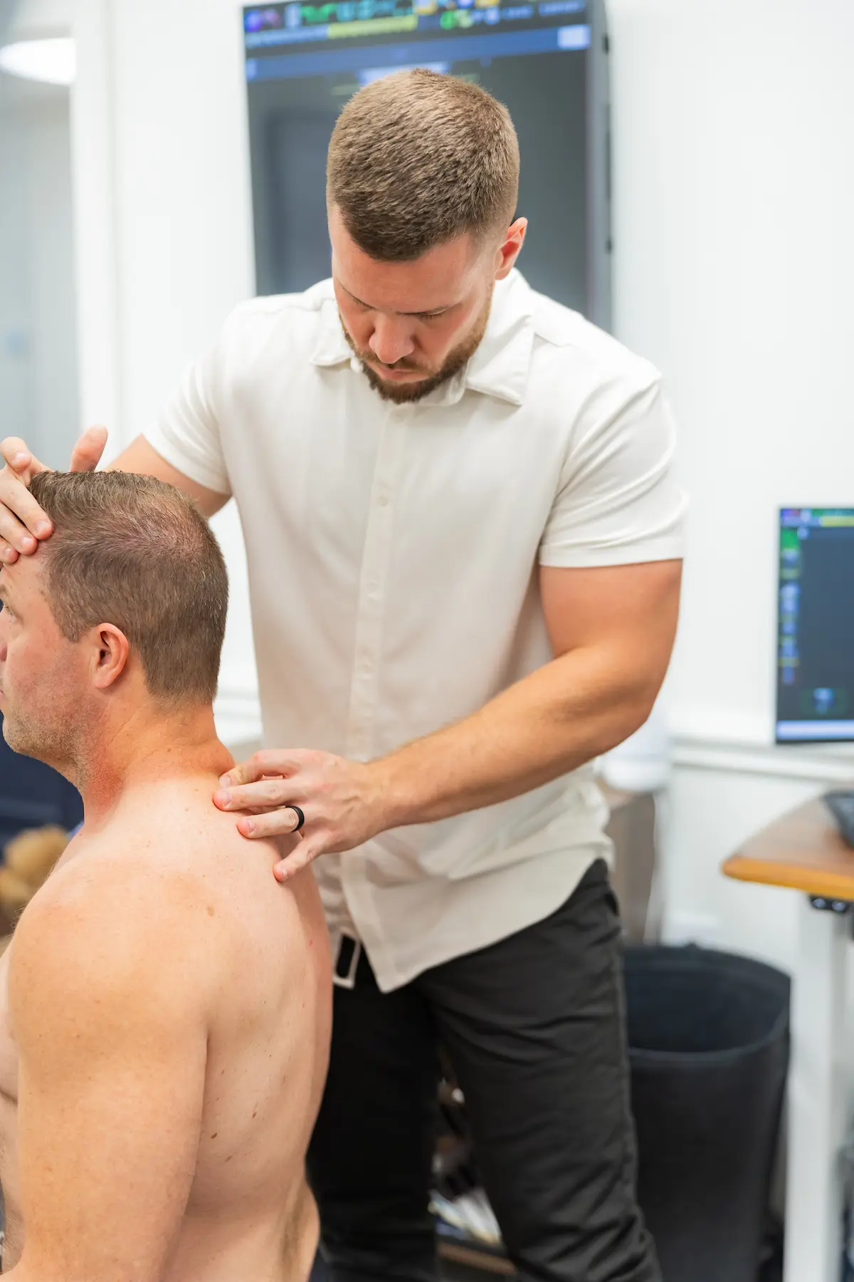 A practitioner in a white shirt examines the neck and shoulder of a shirtless patient from behind.