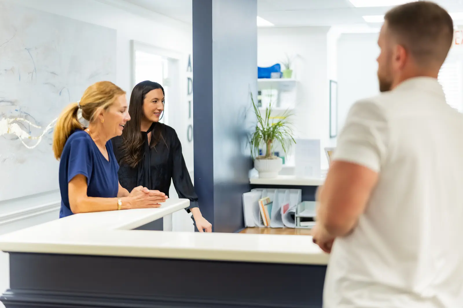 Two women at a reception desk smiling and interacting with a man whose back is to the camera in a modern office setting.