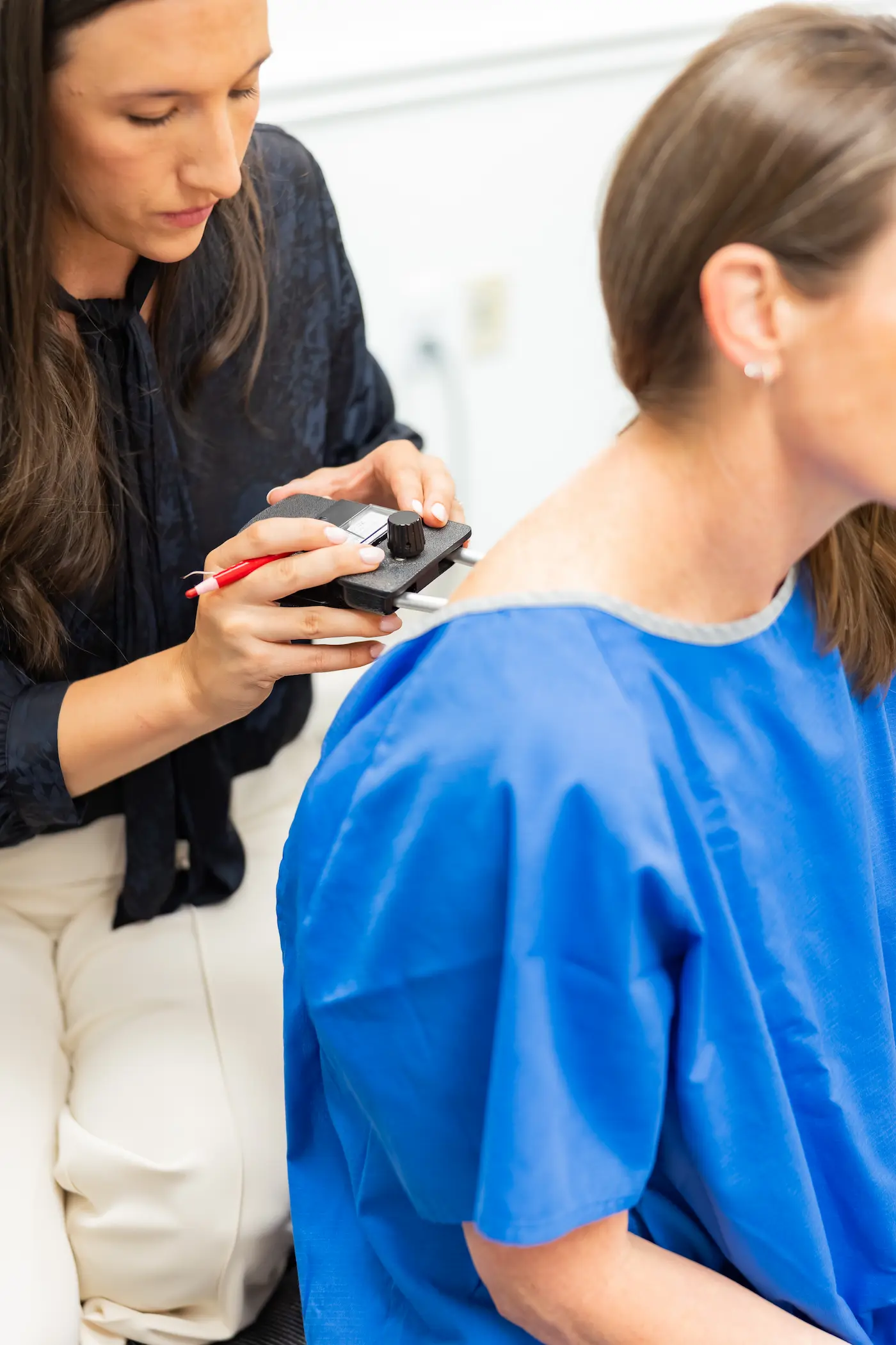 A medical professional examines a patient's shoulder with a black handheld device and a red pen, as the patient wears a blue gown.