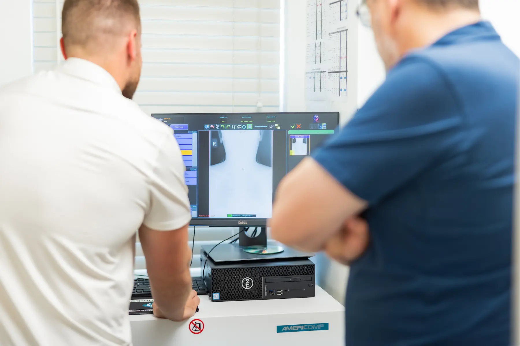 Two men, seen from behind, viewing a neck X-ray image on a Dell computer monitor in a medical office.
