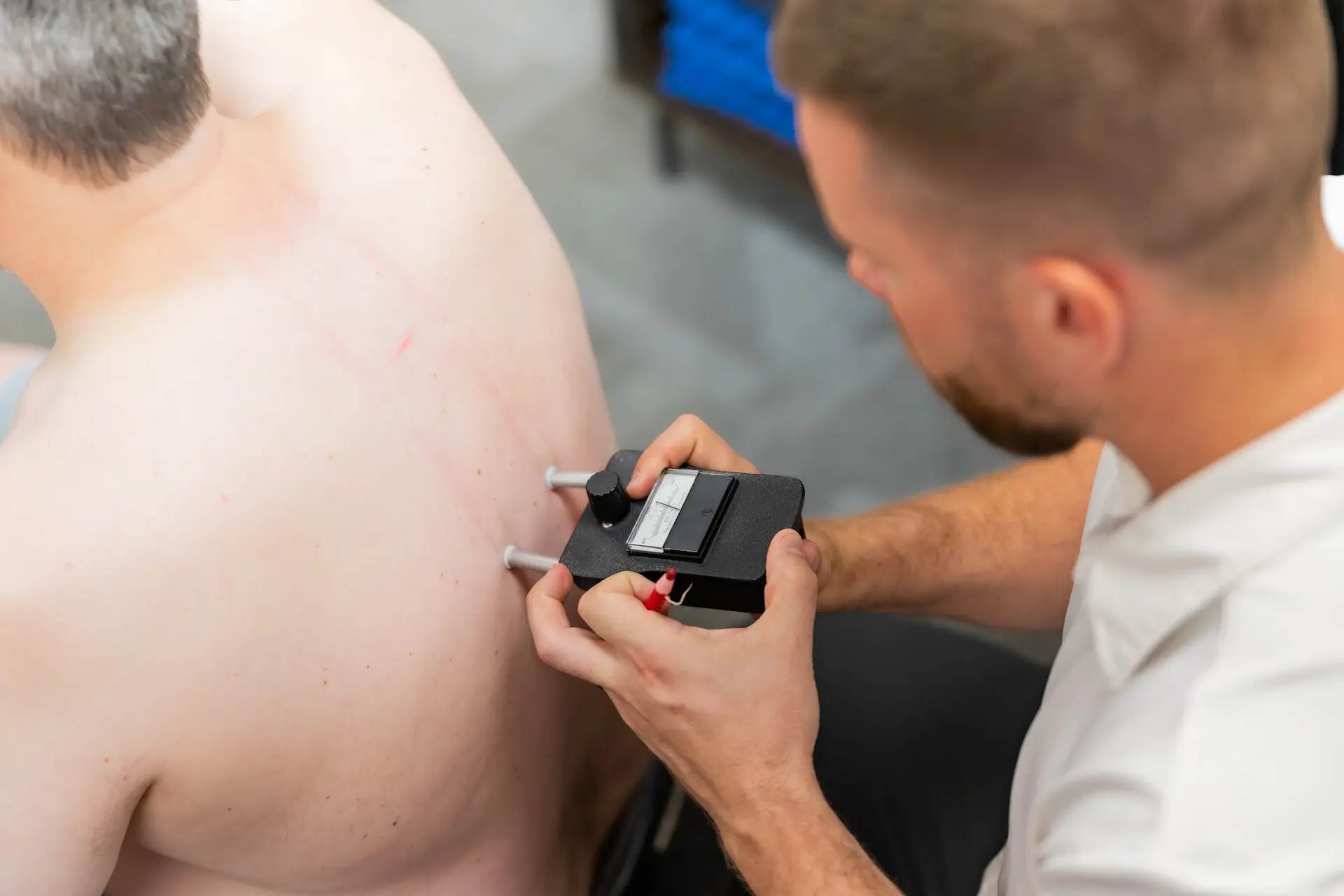A medical practitioner uses a black diagnostic device with probes on a patient's bare upper back.