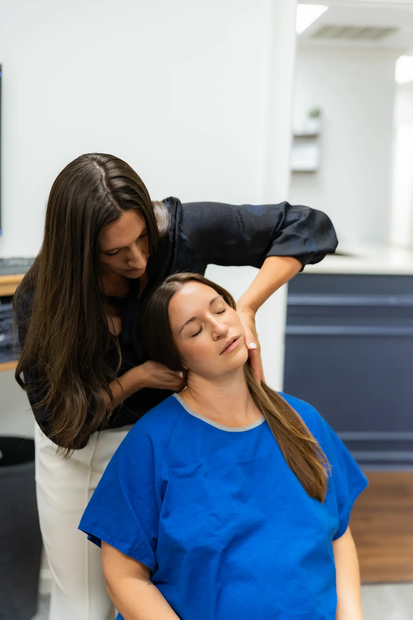 A practitioner with long dark hair adjusts the neck of a patient in a blue gown, whose eyes are closed.
