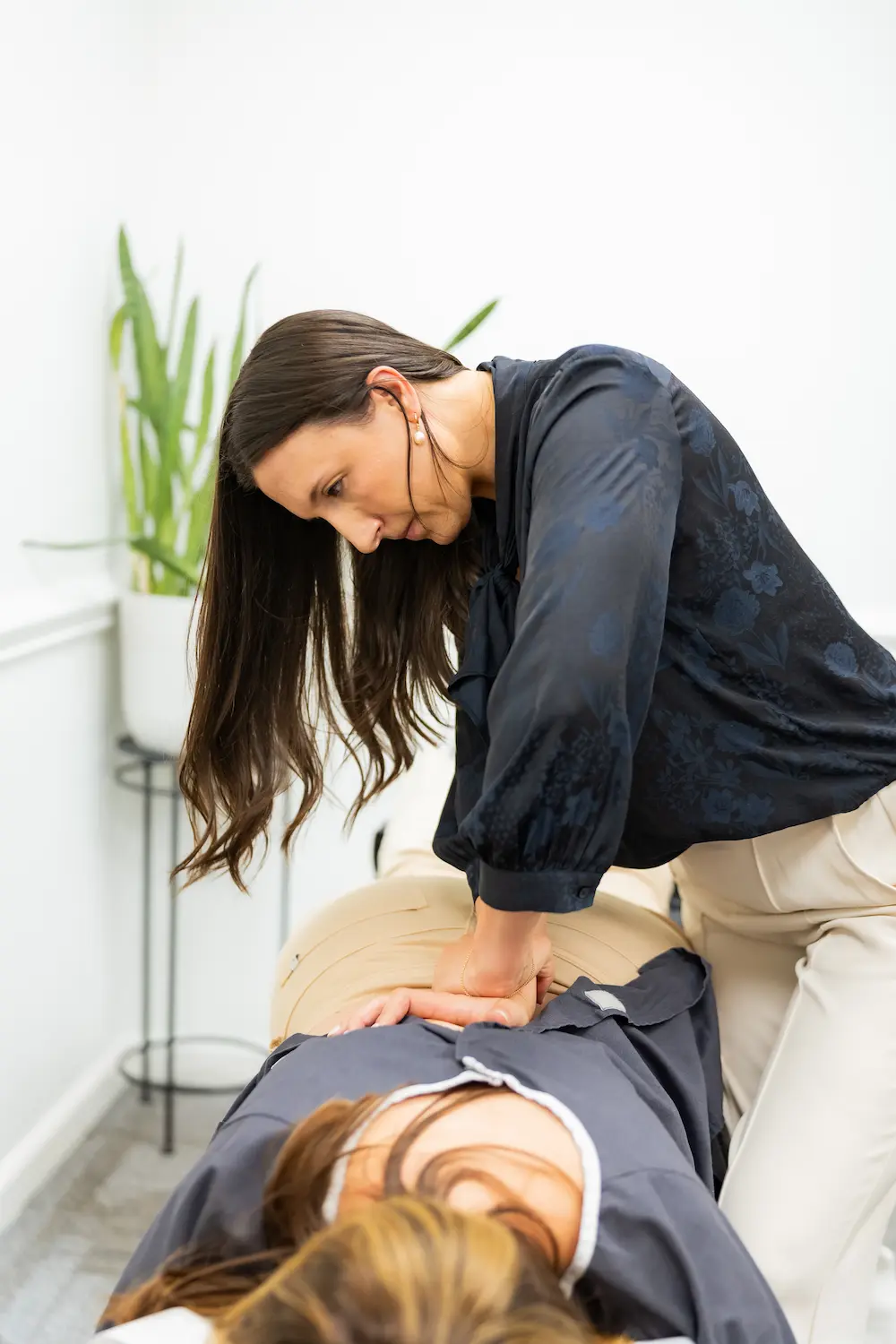 A female chiropractor with dark hair performs a manual adjustment on a patient's lower back, who is lying face down on a table.