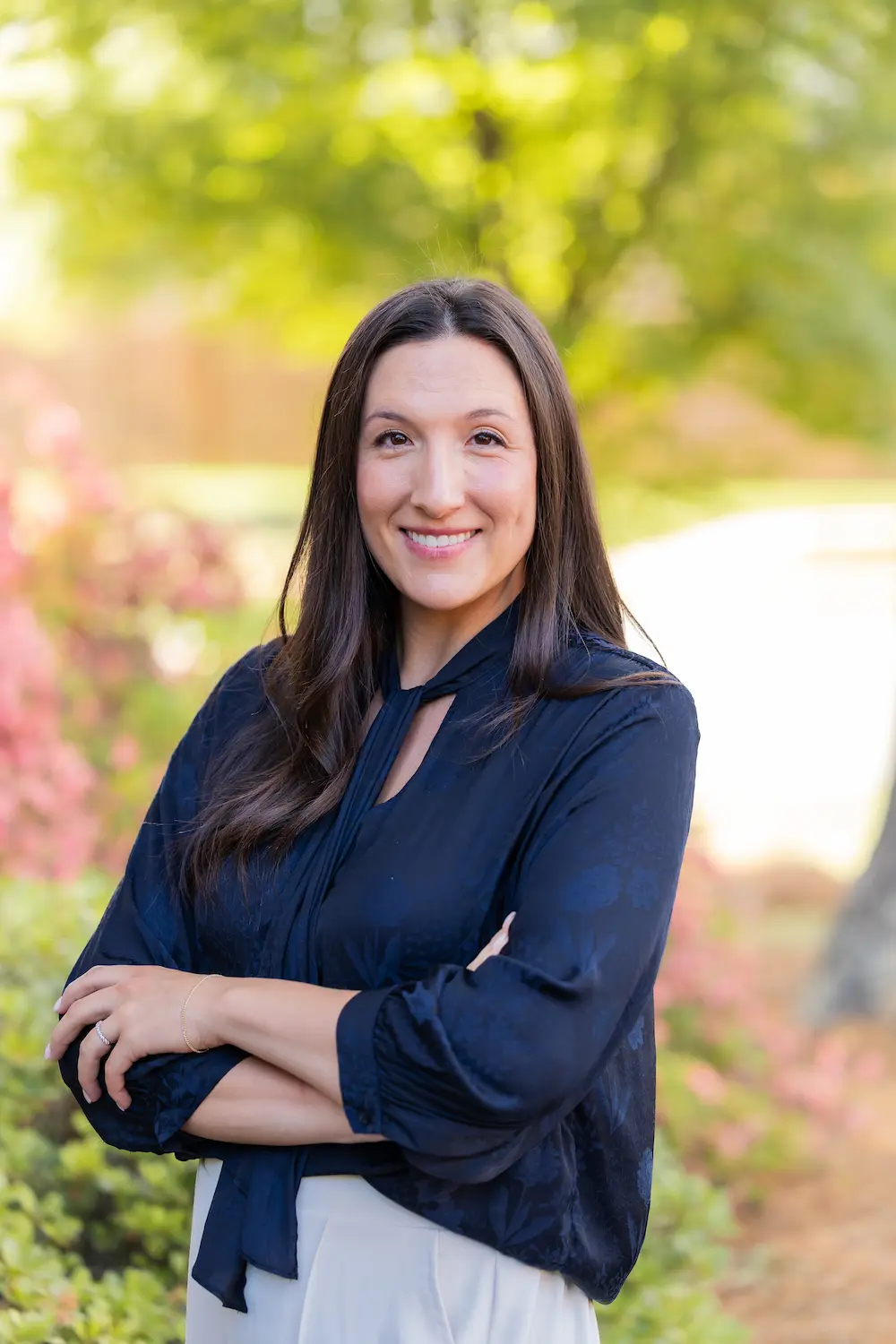 A smiling woman with long dark hair, wearing a navy blue blouse, stands with crossed arms in an outdoor setting with blurred green and pink foliage.