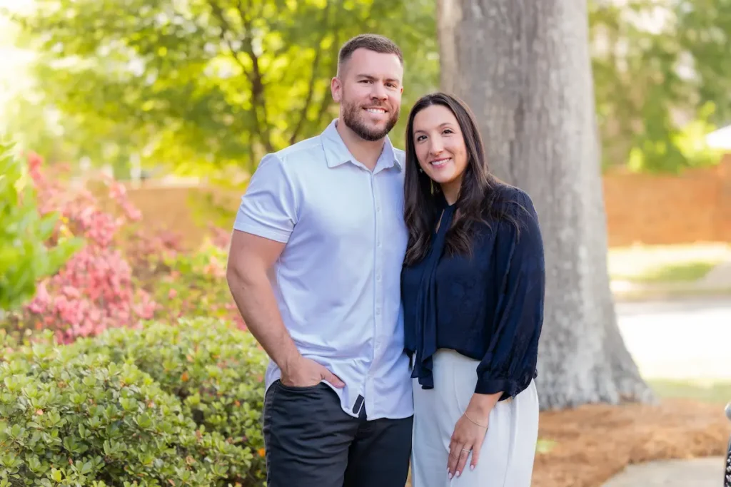 A smiling man and woman standing together outdoors with green plants and a tree in the background.