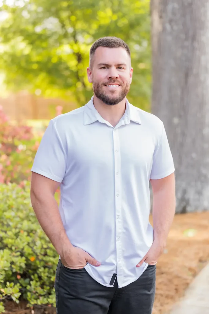 A smiling man with a beard and short dark hair, wearing a white short-sleeved shirt and dark pants, stands with his hands in his pockets outdoors.