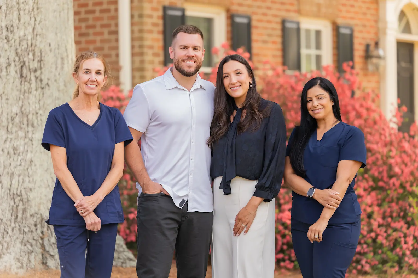 Four smiling individuals, two women in blue scrubs, a man in a white shirt, and a woman in a black blouse, standing outdoors in front of a brick building and red bushes.
