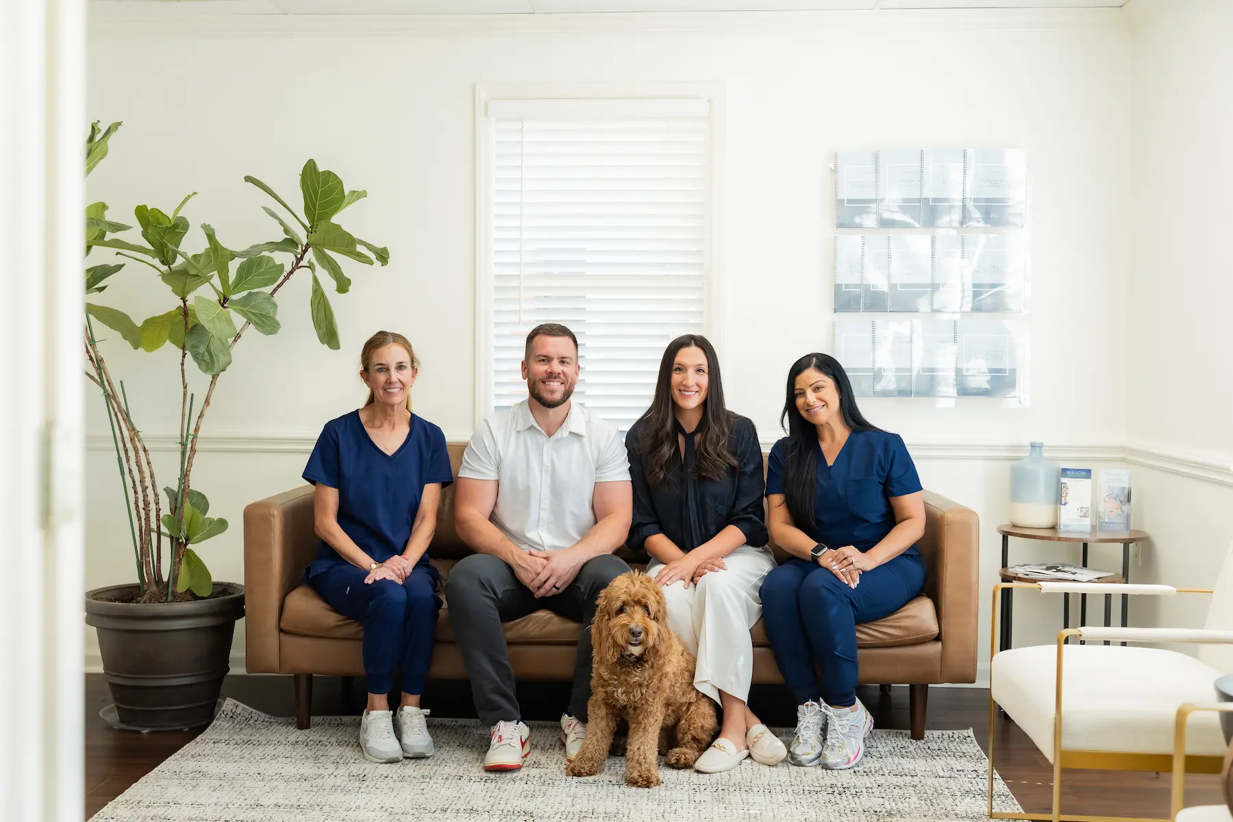 Four smiling people and a brown curly-haired dog sit on a brown couch in a bright office waiting room.